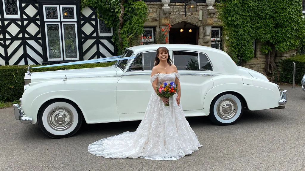 Smiling bride holding a bouquet while standing next to a white Rolls-Royce Silver Cloud