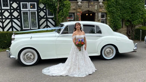 Smiling bride holding a bouquet while standing next to a white Rolls-Royce Silver Cloud