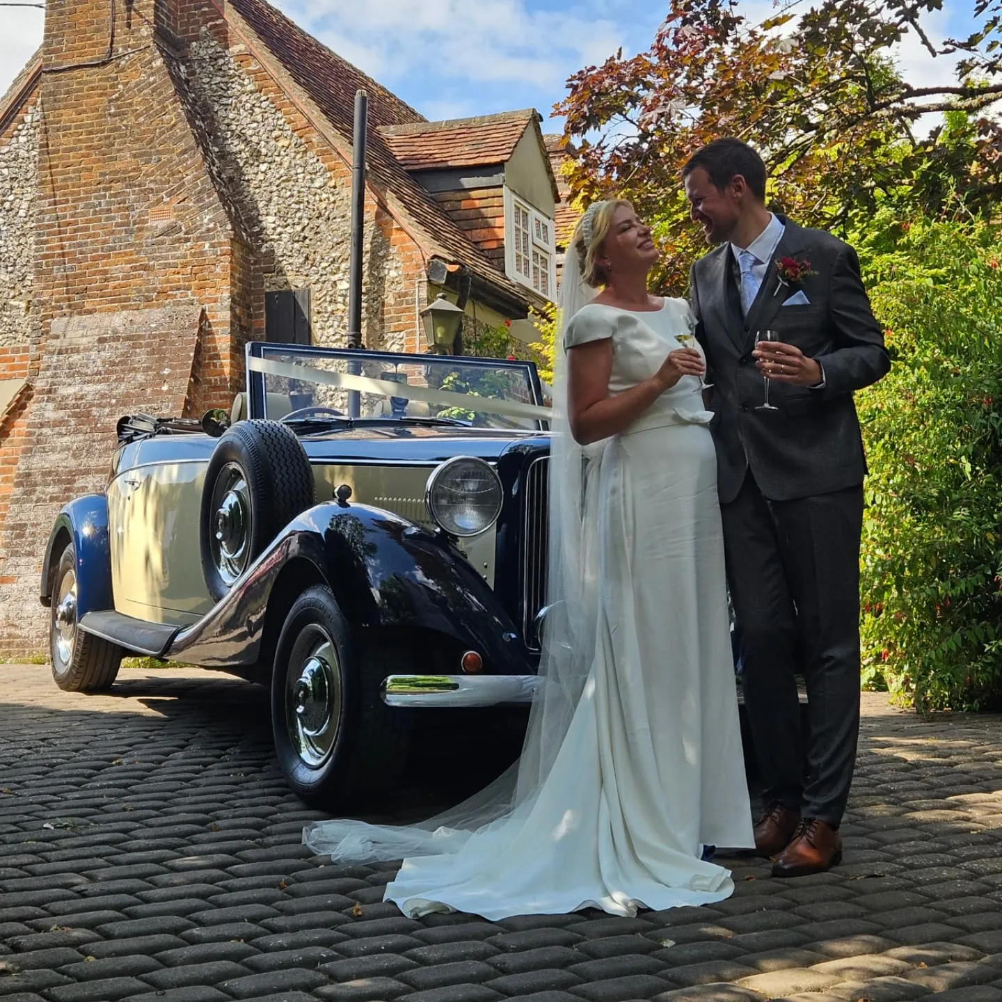 Bride and groom share a ktoast with a glass of Champagne in their hands beside a vintage blue and cream convertible wedding car in a picturesque village setting.