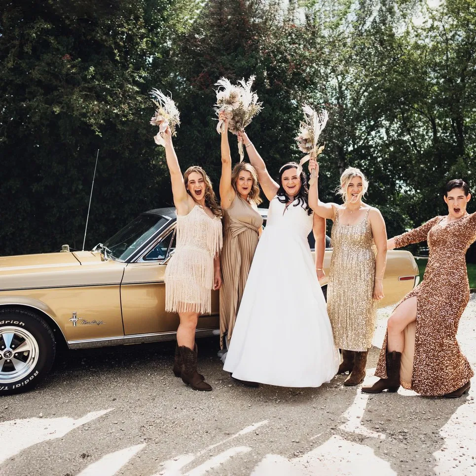 Bride with bridesmaids in vintage-style dresses posing beside a gold American Mustang wedding car.
