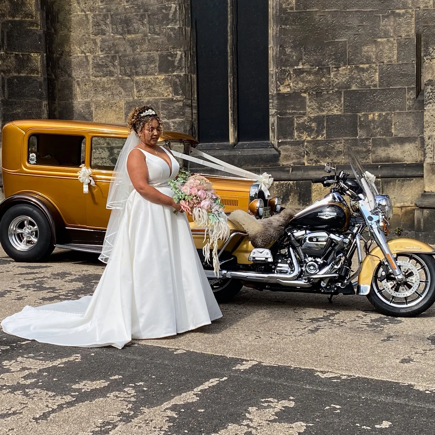 Bride in white wedding gown standing with two unique gold vehicles, including a car and motorcycle, outside a church.