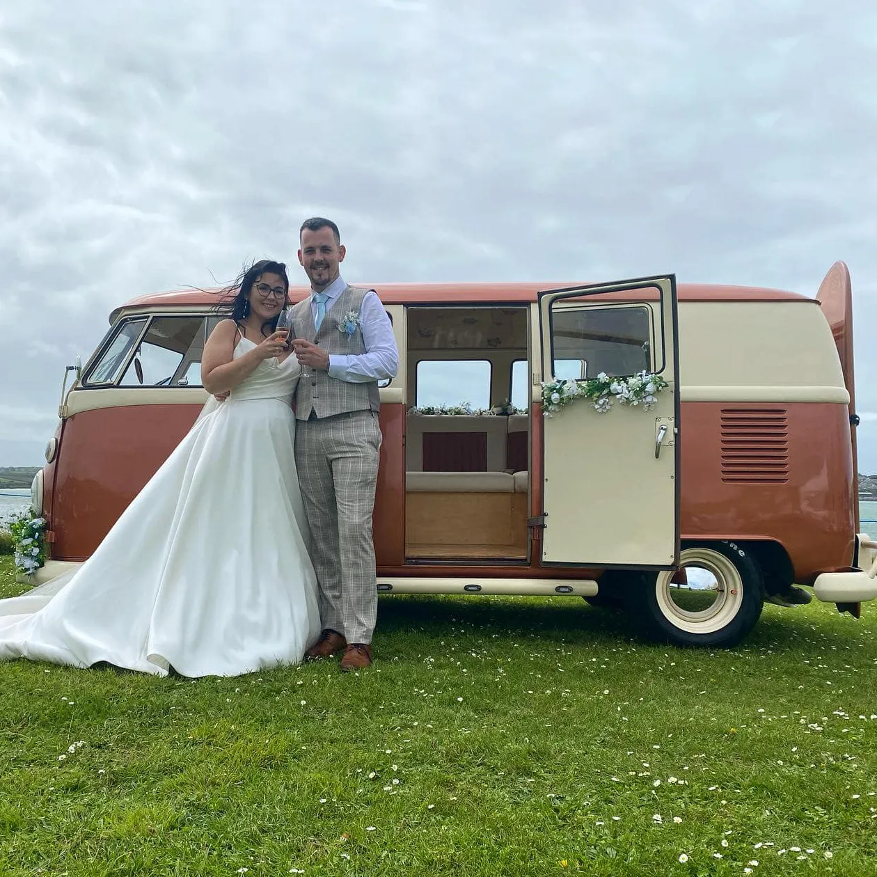Bride and groom embracing beside a burnt orange VW campervan wedding car.