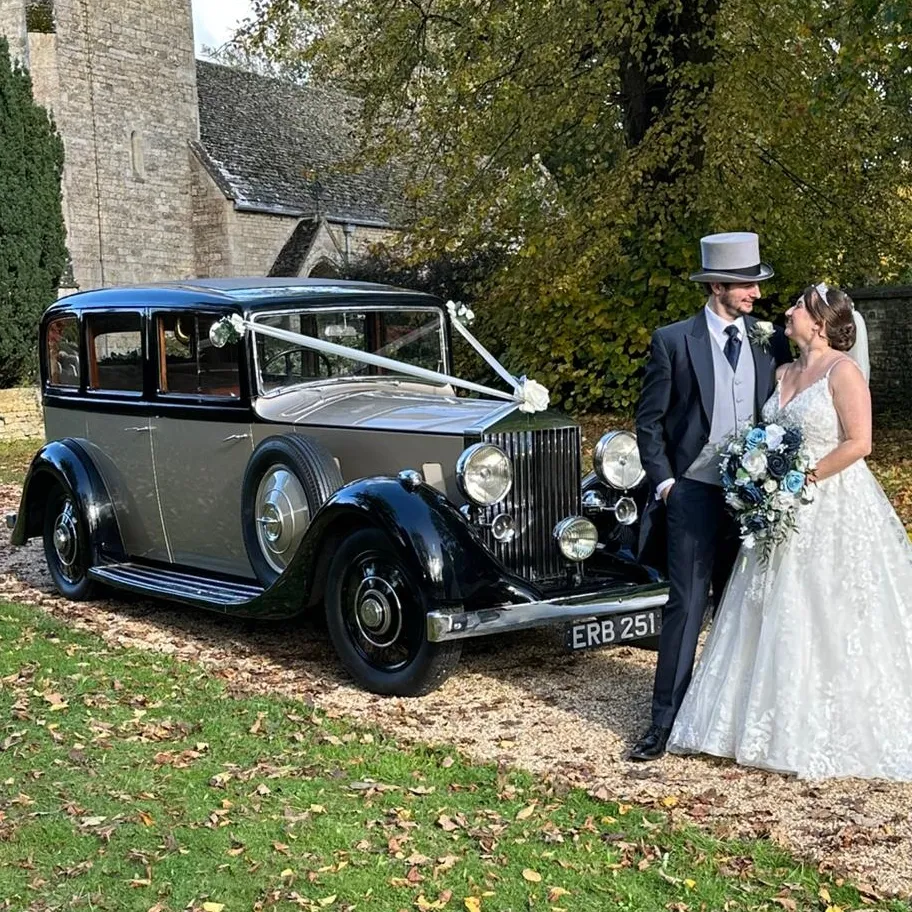 Newly married couple with a champagne-coloured vintage Rolls-Royce wedding car parked outside a church.