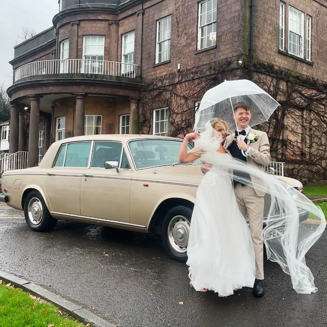 Bride and groom under an umbrella standing beside a gold Rolls-Royce wedding car on a rainy day.