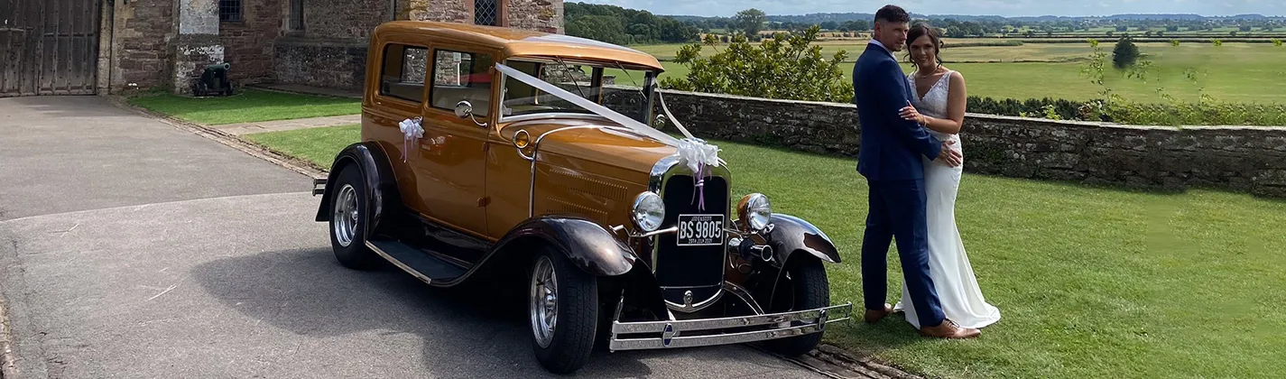 Bride and Groom with a vintage gold wedding car decorated with white ribbons parked on a country estate driveway.