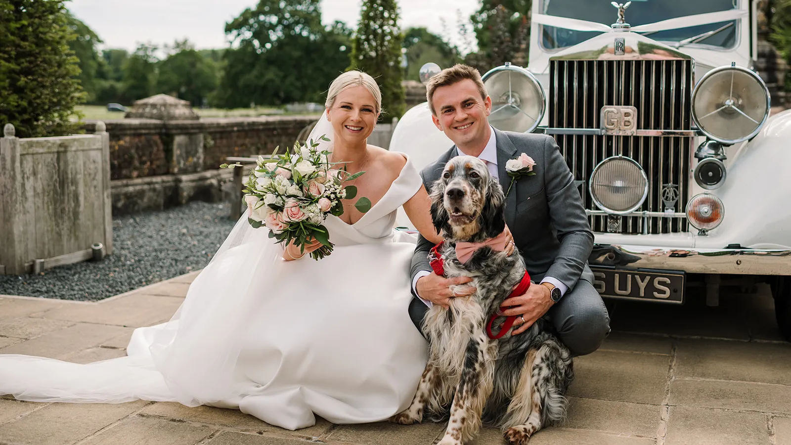 Bride and groom smiling with their dog in front of a classic white Rolls-Royce wedding car. The bride holds a bouquet of pink and white flowers while the dog wears a bow tie, making a charming family wedding portrait.