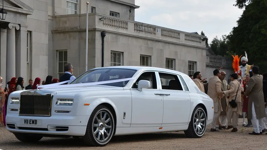Front left angle view of white Rolls-Royce Phantom with Asian groom in the background surrounded by his guests