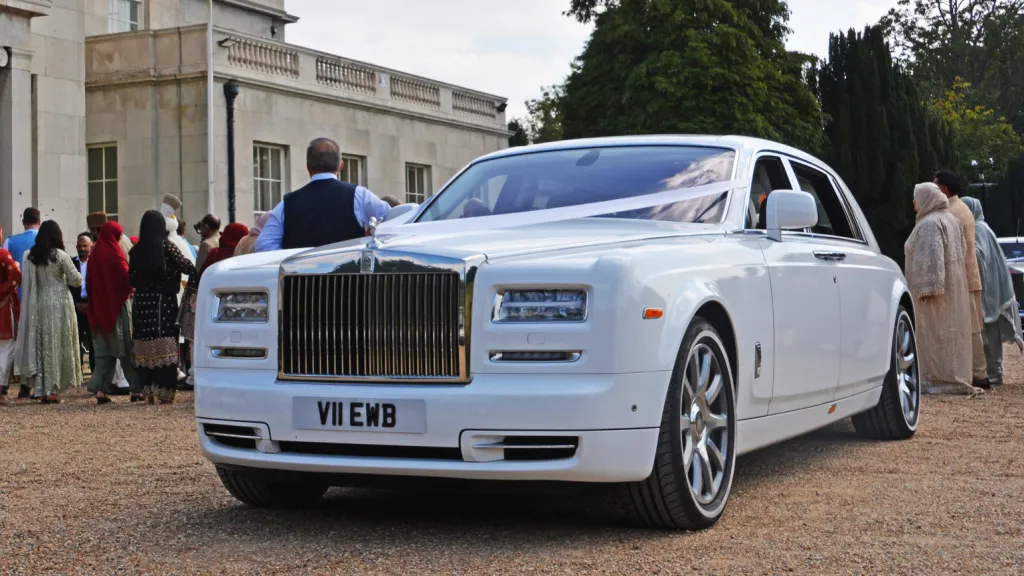 right left angle view of White rolls-royce phantom with large chroime grill, white ribbons and thin white wall tires. Wedding guests can be seen in the background
