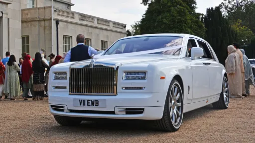 right left angle view of White rolls-royce phantom with large chroime grill, white ribbons and thin white wall tires. Wedding guests can be seen in the background