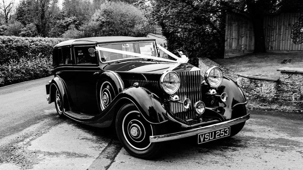 Black and white photograph of a vintage Rolls-Royce parked outside a historic wedding venue, headlights lit and classic chrome details gleaming.