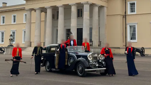 Deep blue vintage Rolls-Royce parked in front of a grand ceremonial building with tall pillars and uniformed guards standing nearby.