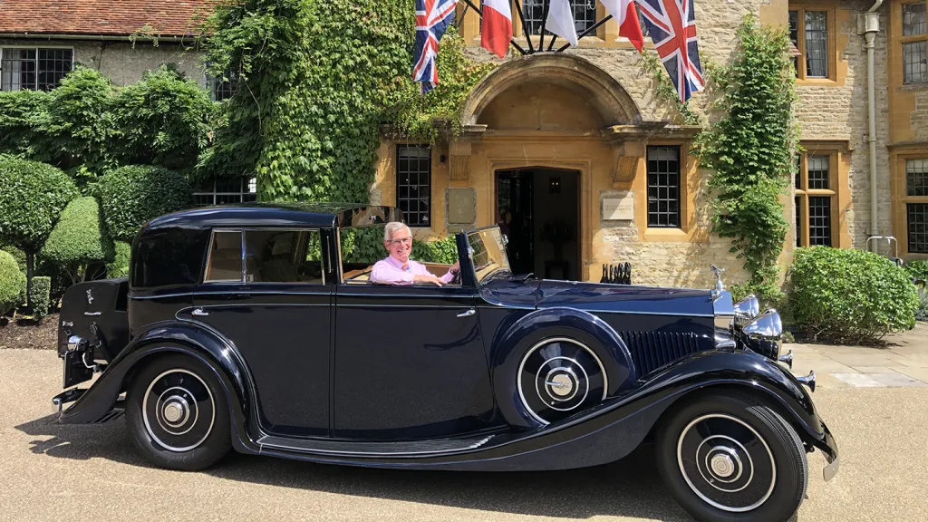 Blue vintage Rolls-Royce with a smartly dressed chauffeur at the wheel, parked outside an elegant building with flags above the entrance.