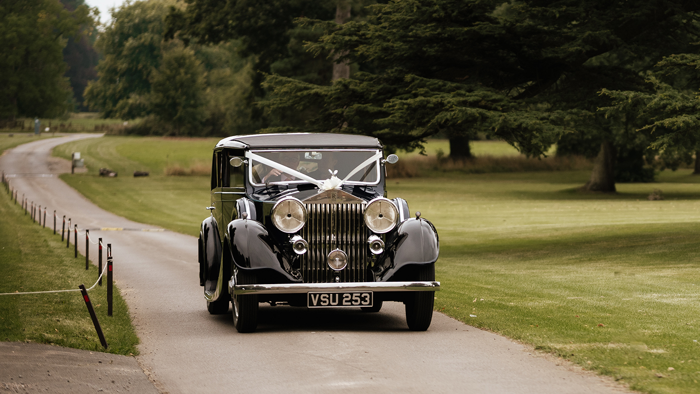 Blue vintage Rolls-Royce decorated with white wedding ribbons, driving up a long country driveway lined with grass and trees.