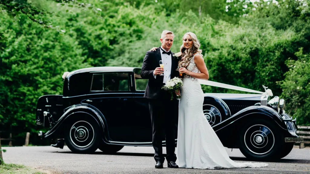 Bride and groom posing together in front of a black vintage Rolls-Royce on a sunny lawn with trees in the background.