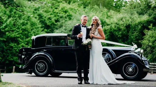 Bride and groom posing together in front of a black vintage Rolls-Royce on a sunny lawn with trees in the background.