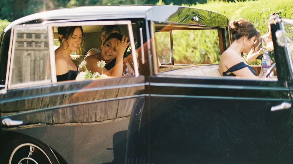 Bride and Bridesmaids seated inside a blue vintage Rolls-Royce, smiling through the open window as the car drives past.