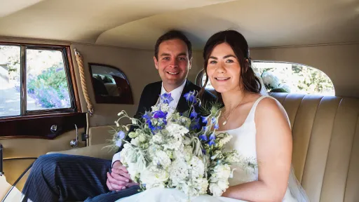 Bride and groom smiling together inside a cream leather interior of a vintage Rolls-Royce, the bride holding a bouquet of white and lilac flowers.
