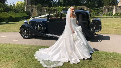 Bride wearing a flowing white gown standing next to a deep blue vintage Rolls-Royce with the long train of her dress spread across the driveway.
