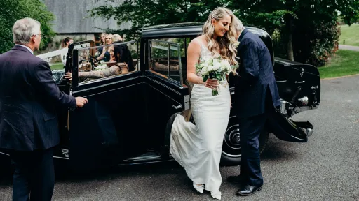 Bride stepping out of a vintage Rolls-Royce as her father offers his hand, the blue car parked on a quiet road and chauffeur is opening door to Bridesmaid seating at the front.