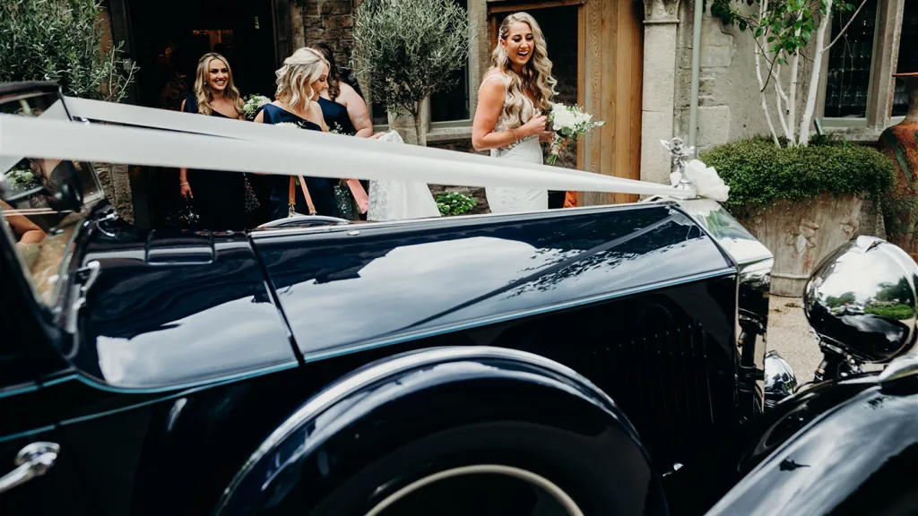 Close-up of white wedding ribbons and bow draped across the bonnet of a blue vintage Rolls-Royce, with bridesmaids standing in the background.