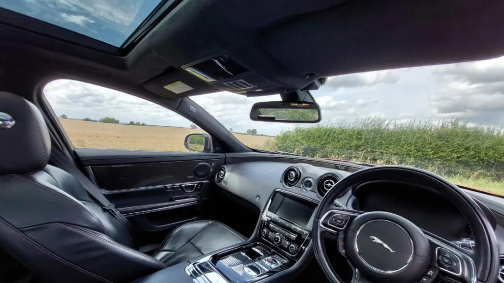 Wide front interior view of a Jaguar XJR, highlighting the sleek black leather seats, centre console and steering wheel against an outdoor backdrop.