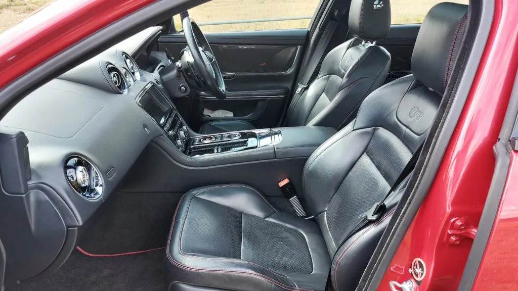 Front passenger and driver seats inside a Jaguar XJR, upholstered in smooth black leather with red exterior paint visible around the door frame.