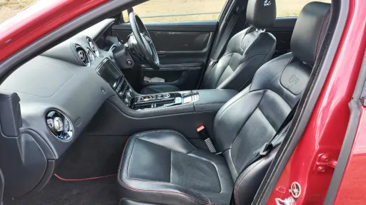 Front passenger and driver seats inside a Jaguar XJR, upholstered in smooth black leather with red exterior paint visible around the door frame.