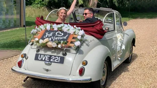 Classic Morris Minor Convertible right side view with roof open and big large ivory bows on door handle and just married sign at the rear with Bride and Groom weaving inside the vehicle