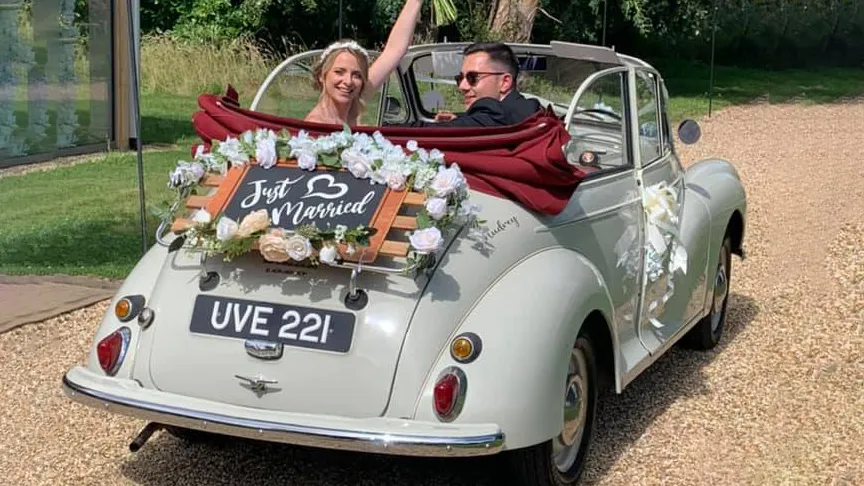 Classic Morris Minor Convertible right side view with roof open and big large ivory bows on door handle and just married sign at the rear with Bride and Groom weaving inside the vehicle