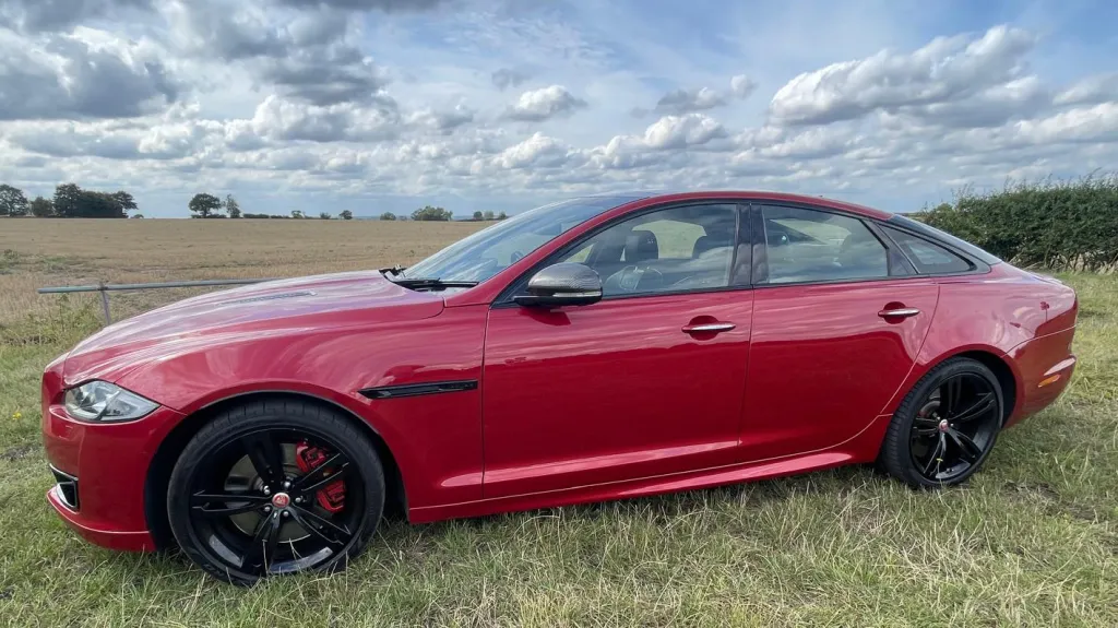 Side profile of a red Jaguar XJR parked in an open field, featuring black alloy wheels and sleek sports styling.
