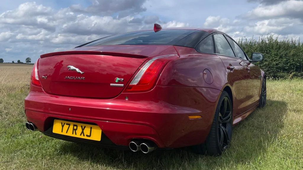Rear exterior view of a red Jaguar XJR with twin exhausts, black wheels and a countryside field in the background.