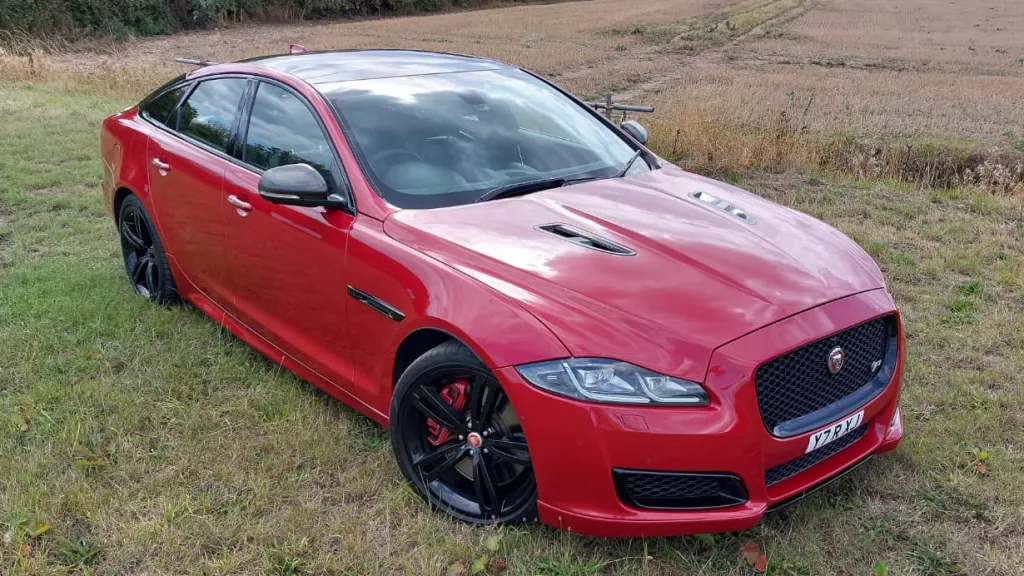 Three-quarter front view of a red Jaguar XJR with striking black alloy wheels, parked on a grassy field.