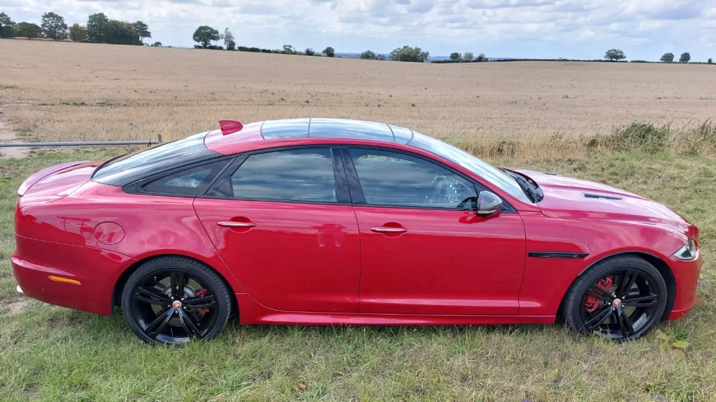 Right side profile of a red Jaguar XJR parked in the countryside, with tinted windows and black alloy wheels.
