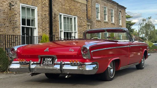 Rear view of a red classic Dodge convertible parked in front of a Georgian-style brick building, showing chrome bumper and tailfins.