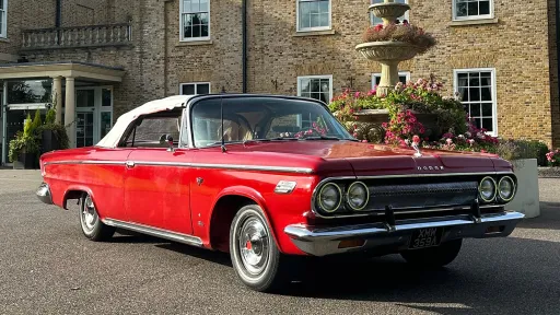 Front angled view of a red Dodge classic convertible with white soft-top roof, parked outside a traditional brick building.