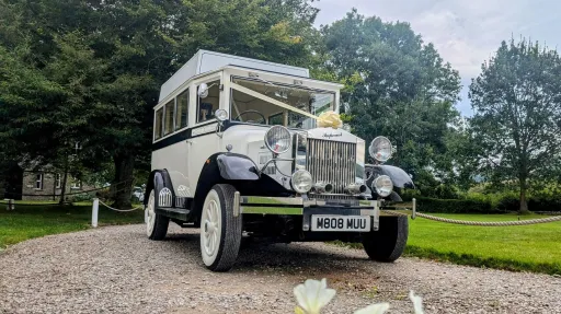 Front view of a white Imperial vintage wedding bus with chrome grille and headlights, parked on a gravel driveway with trees behind.