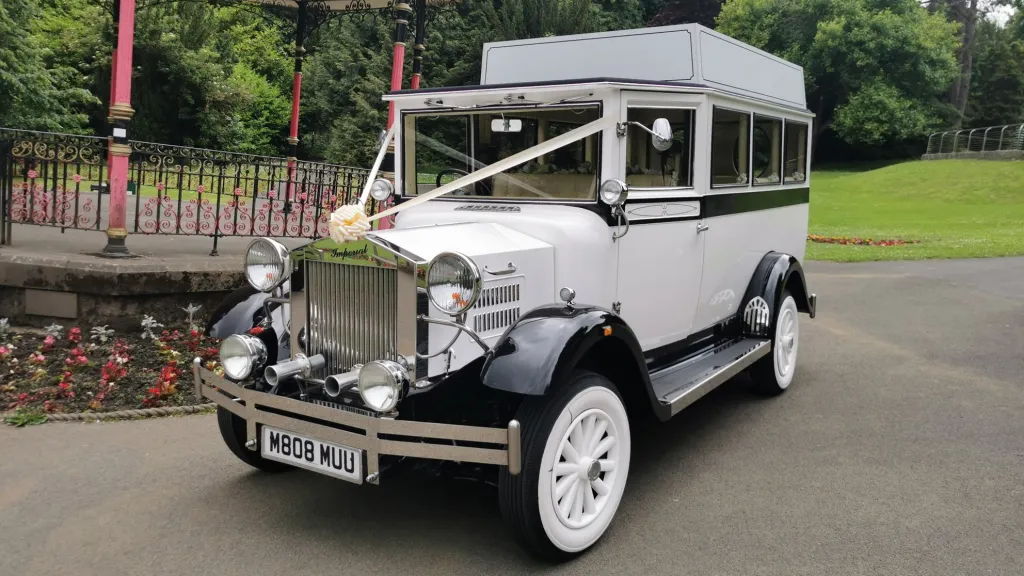 White Imperial vintage wedding bus decorated with white ribbons, parked in front of a landscaped garden and stone walls.