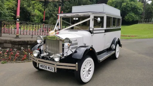 White Imperial vintage wedding bus decorated with white ribbons, parked in front of a landscaped garden and stone walls.
