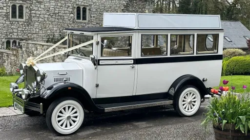 Side profile of a white Imperial vintage wedding bus with chrome headlights and white wheels, parked in front of a stone building.