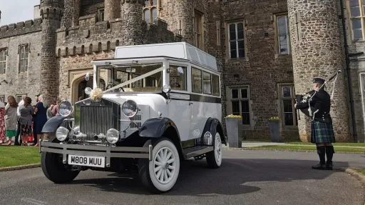White Imperial vintage wedding bus parked at a wedding venue with a bagpipe player standing beside the vehicle