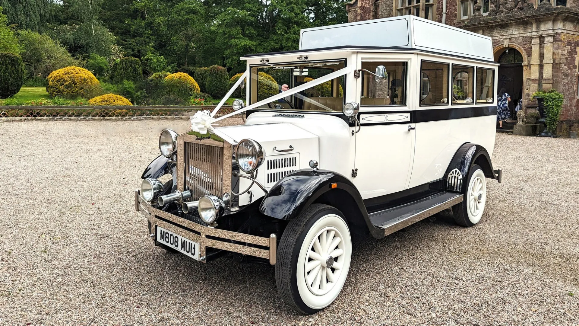 White and black Imperial vintage wedding bus parked on a driveway in Norfolk with greenery in the background.