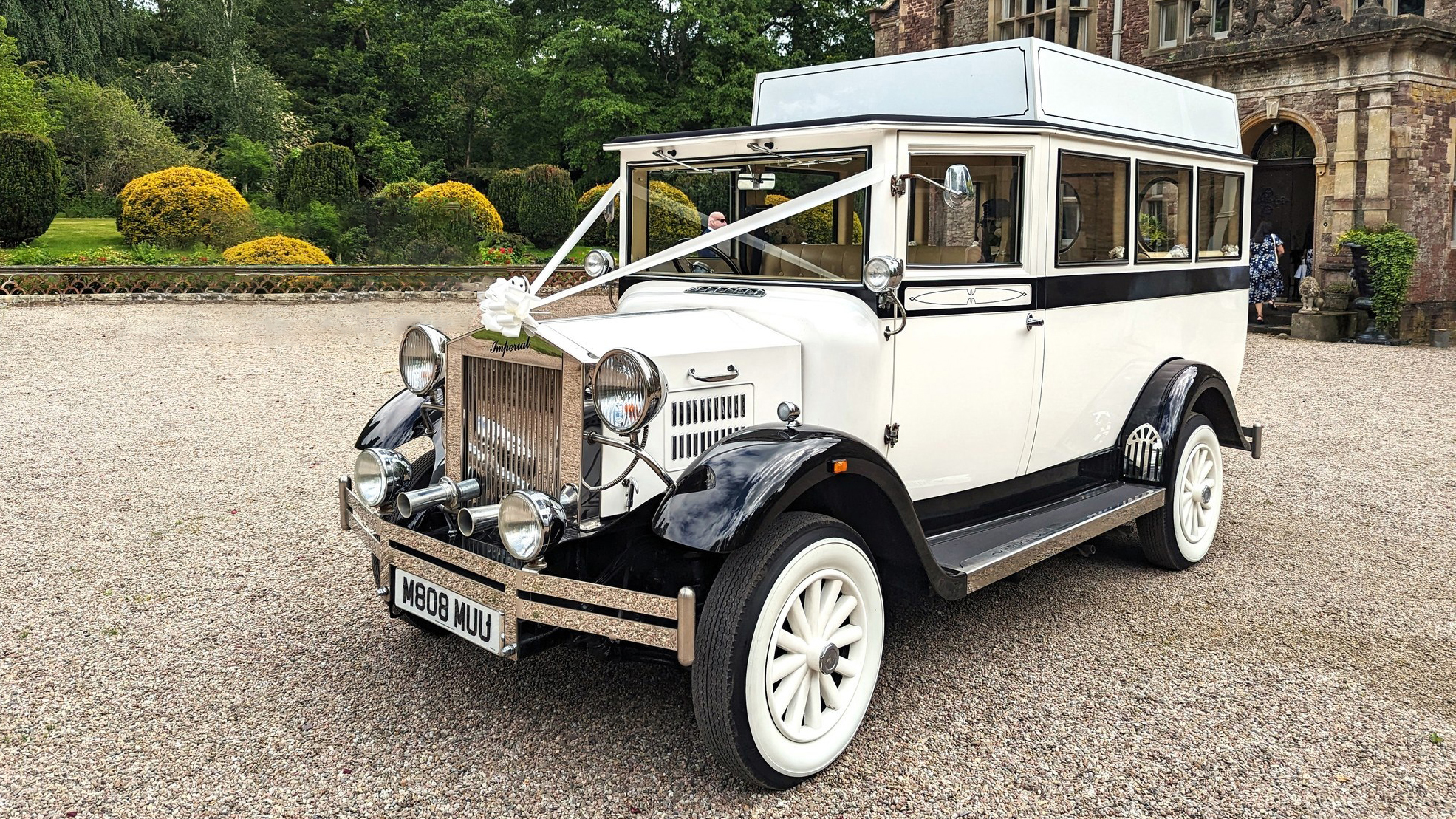 White and black Imperial vintage wedding bus parked on a driveway in Norfolk with greenery in the background.