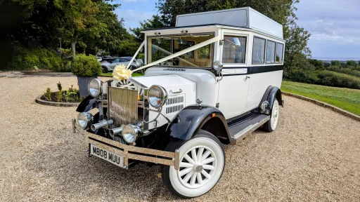 Front angled view of a white Imperial vintage wedding bus with ribbons, parked outside a countryside wedding venue.