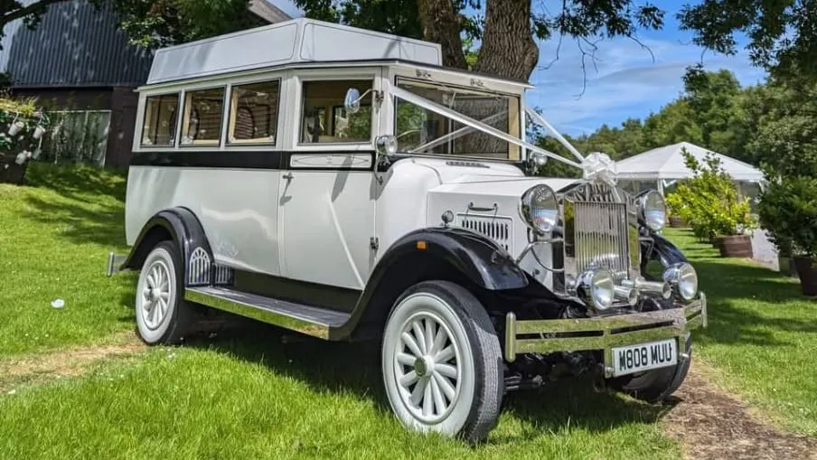 Side and front angled view of a white Imperial vintage wedding bus with ribbons, parked near a stone wedding venue.