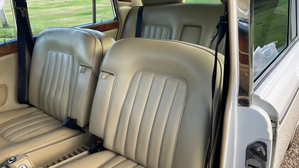 Front interior of a Rolls-Royce Silver Shadow with cream leather seats and polished wooden trim on the doors.