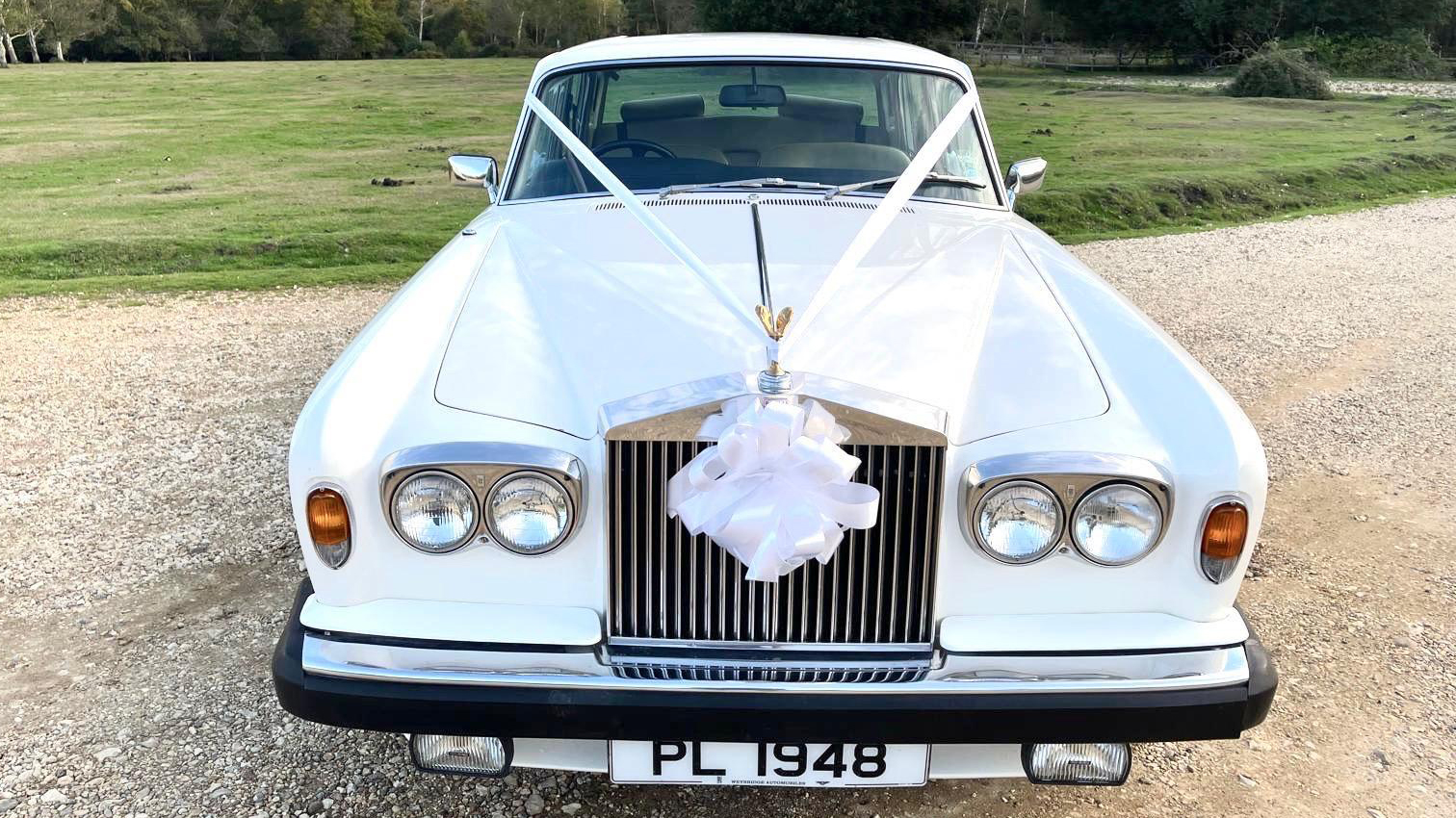 Front view of a white Rolls-Royce Silver Shadow decorated with a large white wedding bow on the grille, parked on a gravel driveway with grass in the background.
