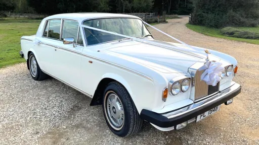 Side angled view of a white Rolls-Royce Silver Shadow with a large white bow on the bonnet, parked outdoors on a gravel driveway in Hampshire.