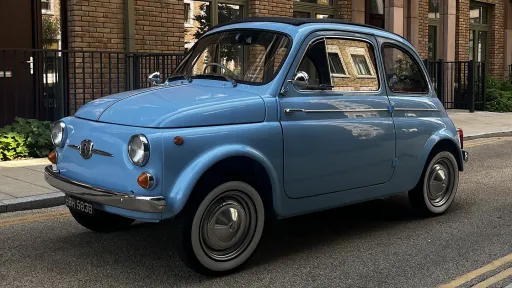 Front angled view of a light blue classic Fiat 500 with whitewall tyres, parked on a quiet London street with brick buildings in the background.
