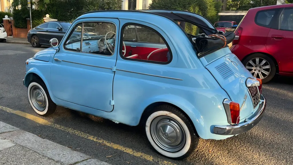 Side profile of a light blue convertible Fiat 500 with chorcoal black fost top roof open, whitewall tyres, parked beside a red car and residential houses.