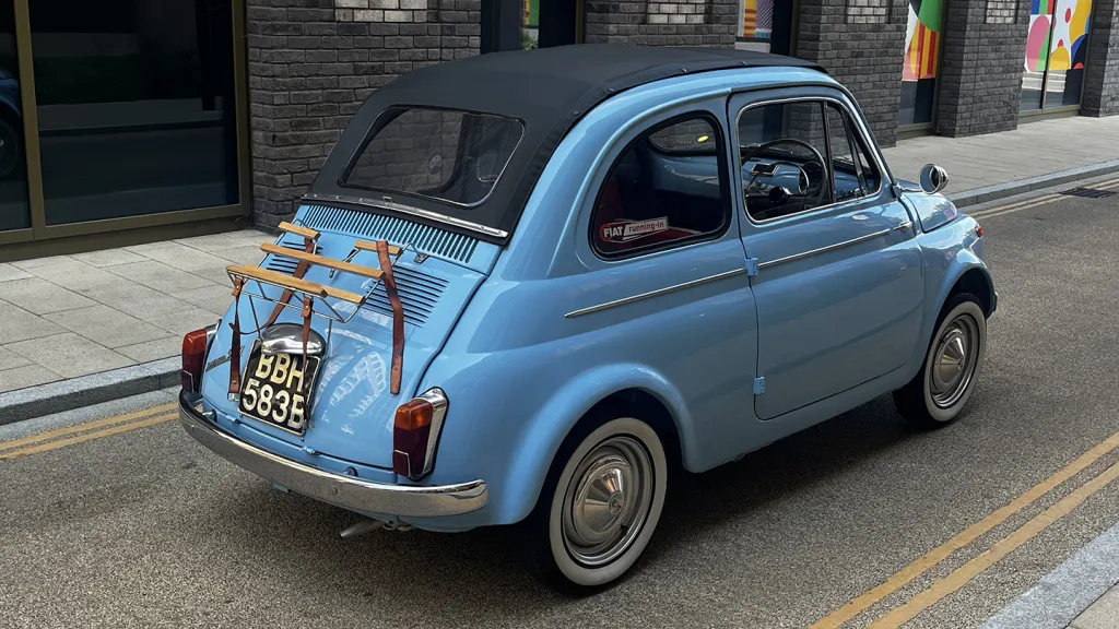 Rear angled view of a light blue Fiat 500 with whitewall tyres, parked on a cobbled street with shops in the background.